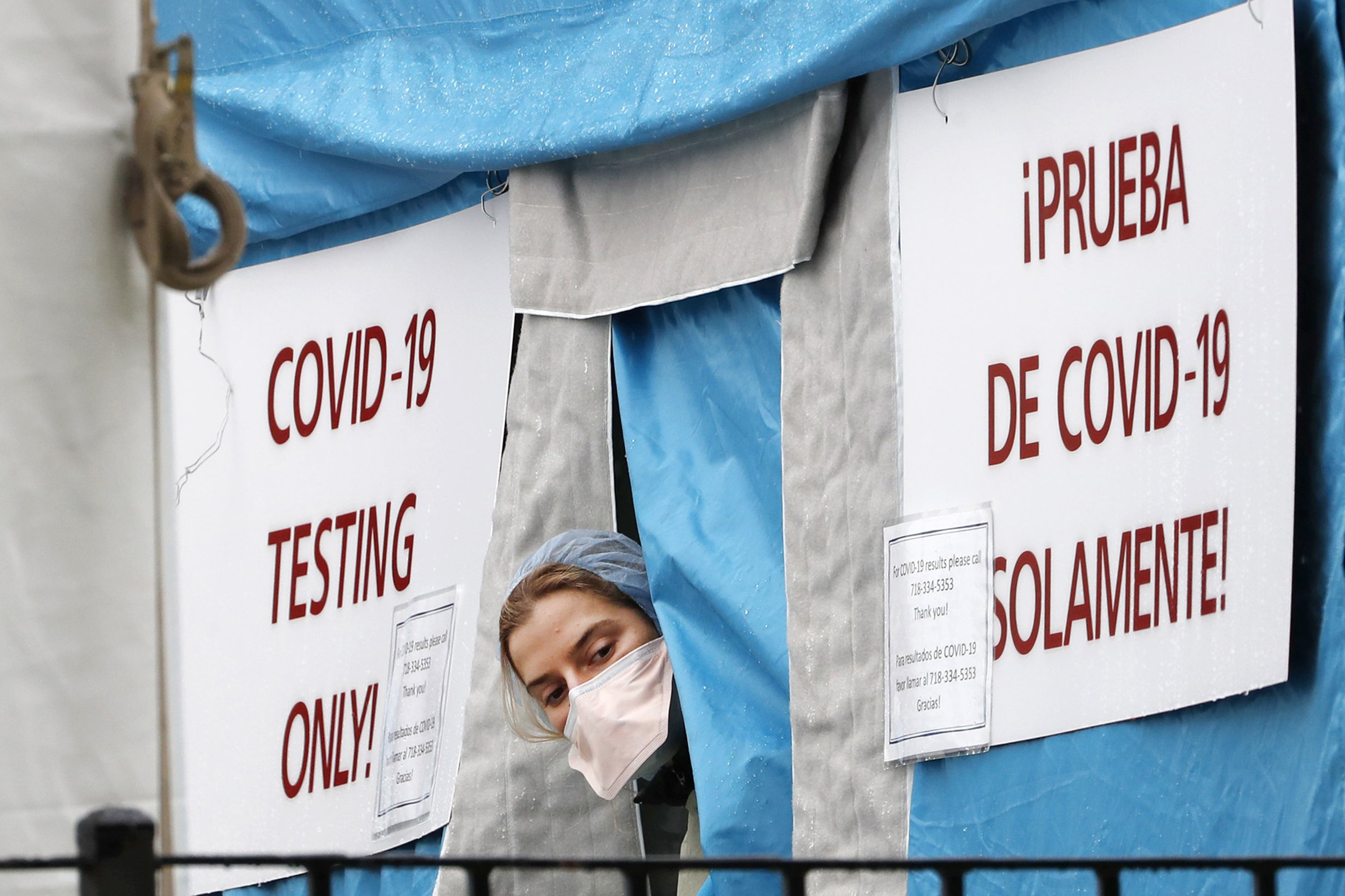 Nurse in scrubs and mask looking out from a temporary tent hung with signs for 'Covid-19 testing' in English and Spanish